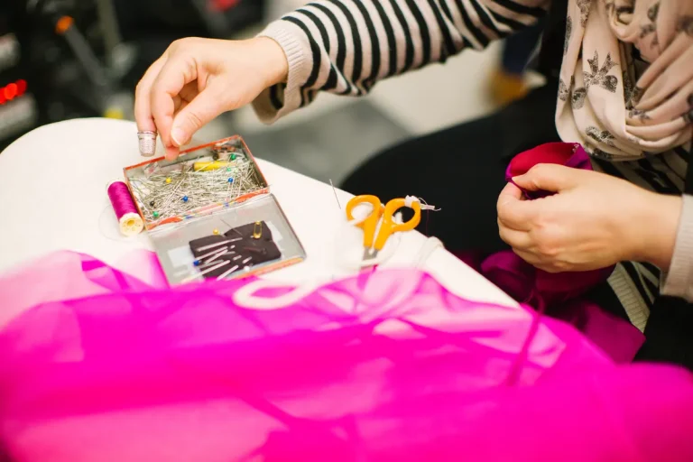 Couturière prenant une épingle dans une boîte, à côté d'une bobine de fil, de ciseaux et d'un tissu fuchsia