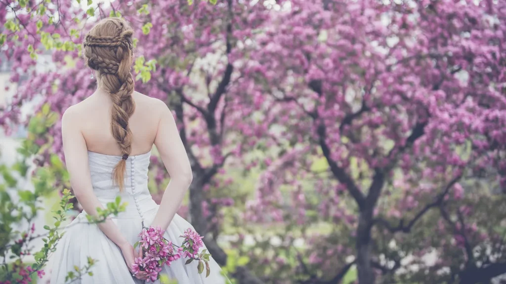 Mariée de dos, longue tresse et bouquet de fleurs roses à la main, sous des arbres en fleurs