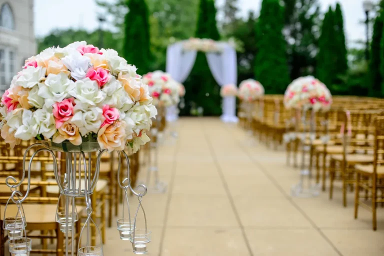Allée de cérémonie de mariage en plein air bordée de bouquets de roses et de chaises dorées, arche en voilage blanc au fond